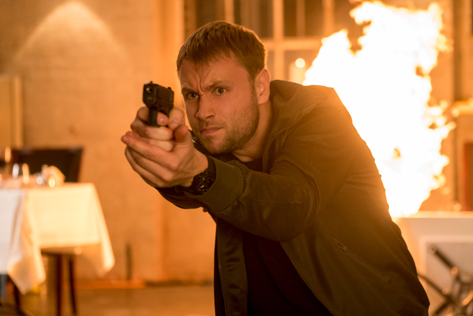 A close up of Max Riemelt holding a gun out, with flames in the background